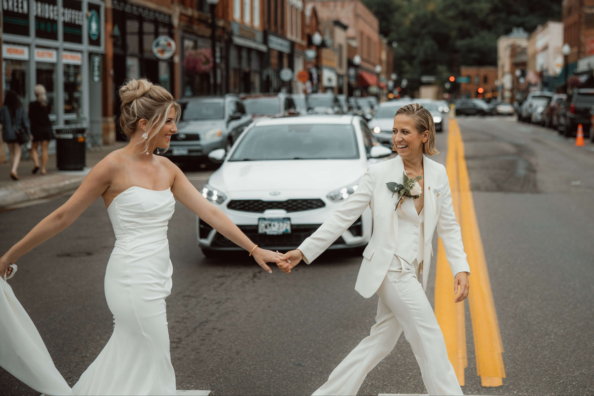A bride in a strapless white wedding gown and a bride in in a white suit holding hands while walking across a city street, with cars and brick buildings in the background.