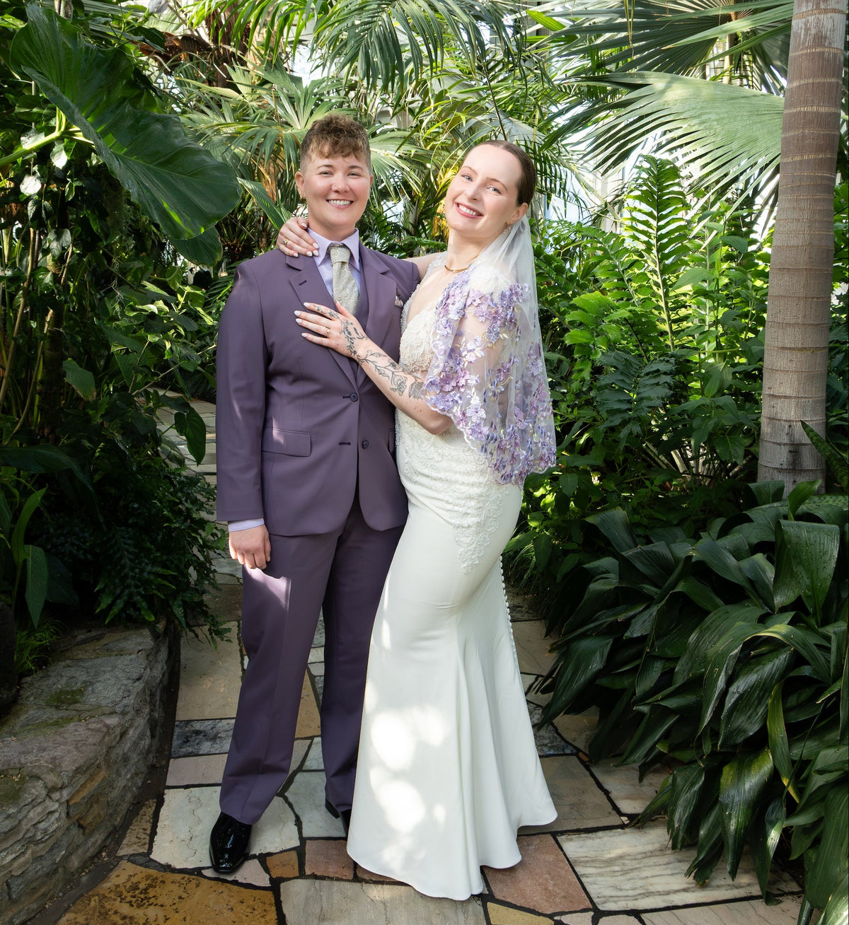 Two brides, one in a white wedding gown and veil and another in a purple suit standing together on a stone path surrounded by lush tropical greenery in a botanical garden.